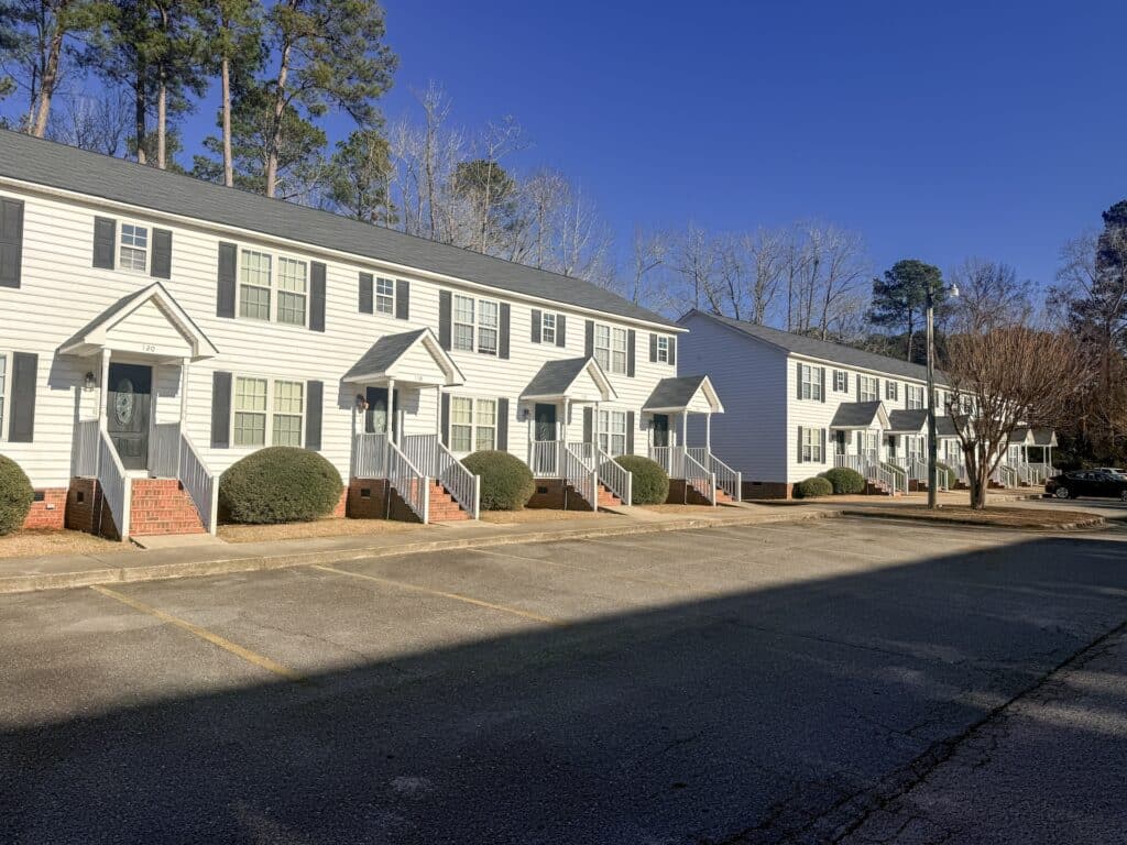 A row of white townhouse-style buildings with brick steps, set against a backdrop of trees under a clear blue sky. The foreground shows an empty parking lot.
