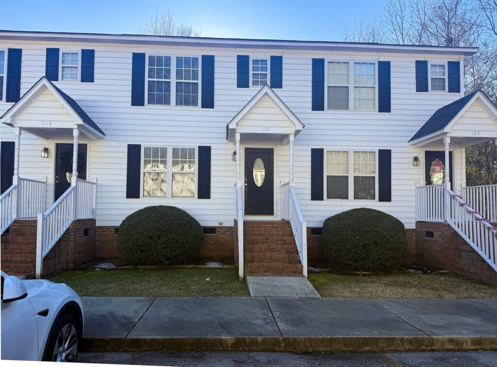 A two-story white townhouse with black shutters, brick steps, and a dark front door. Two symmetrical bushes are in front. A car is parked on the street.