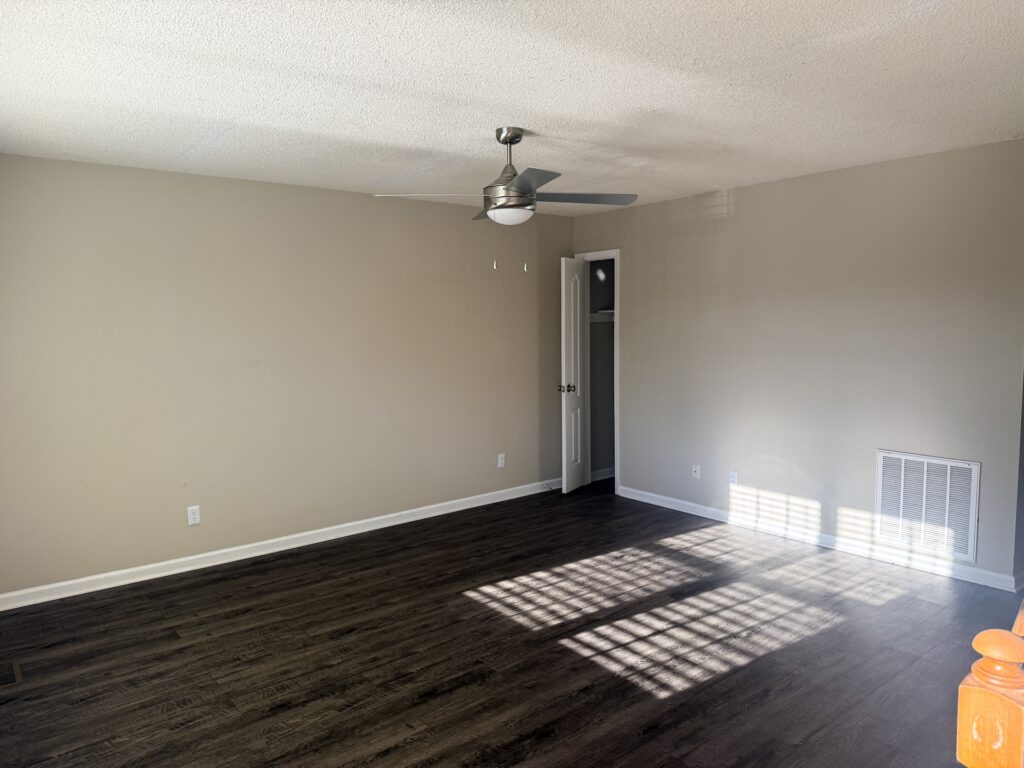 Empty room with beige walls, dark wood floor, a ceiling fan, a closet with white doors, and light streaming through a window casting shadows on the floor.