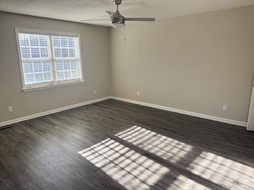 Empty room with gray walls, dark wood flooring, a ceiling fan, and sunlight streaming through a window casting shadows on the floor.