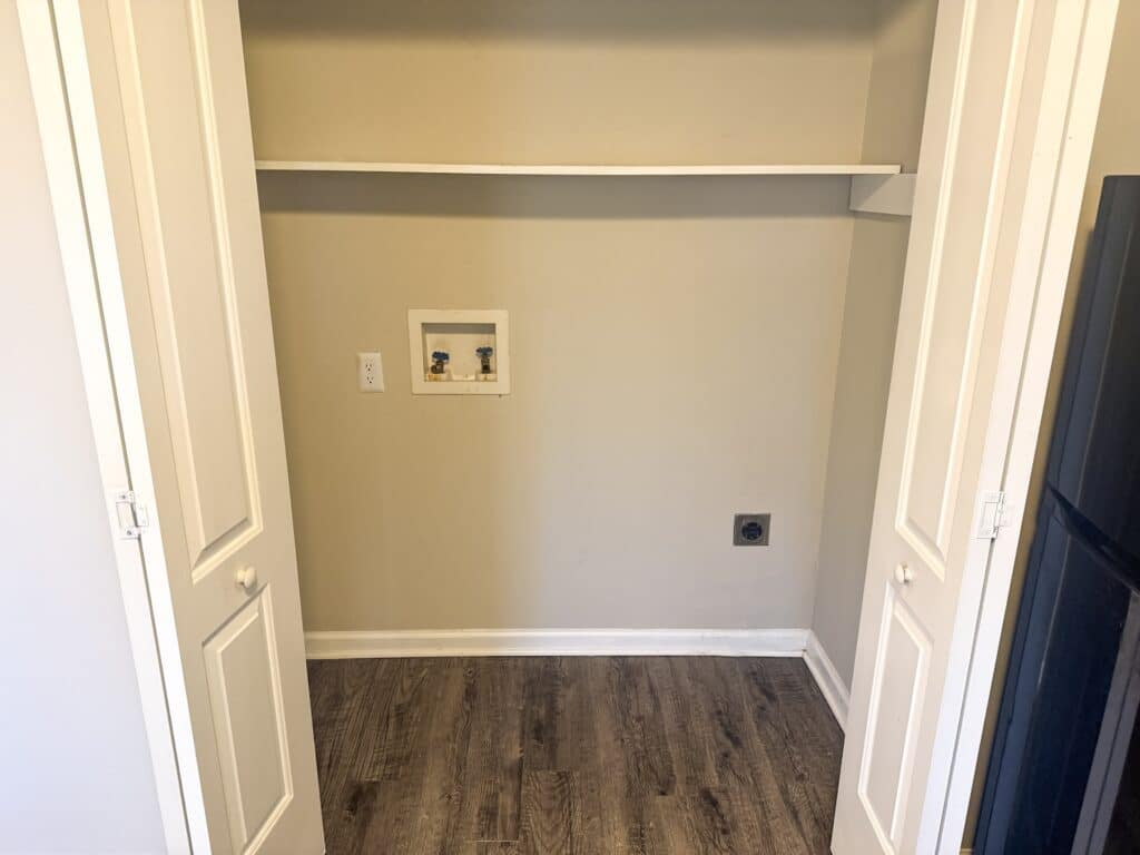 A small, empty laundry nook with wood flooring, beige walls, and a white shelf. It features water connections and a power outlet, flanked by white folding doors.