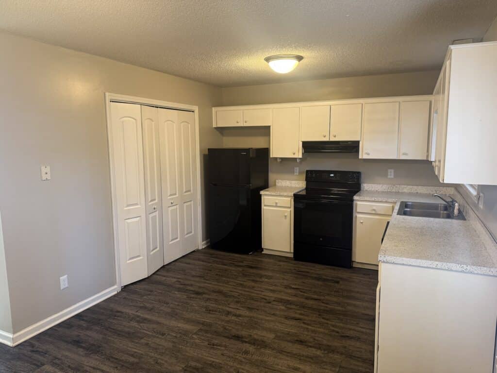 A kitchen with white cabinets, black appliances, and wood flooring. There is a ceiling light and a door to the left.