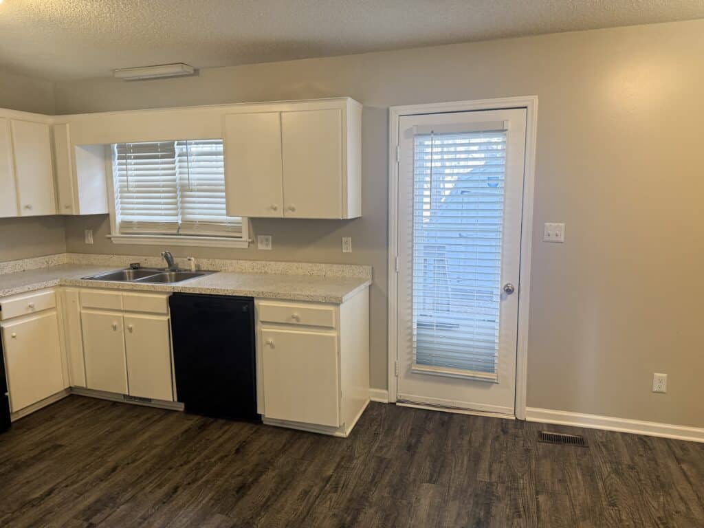 A kitchen with white cabinets, a double sink, and a black dishwasher. There's a window with blinds above the sink and a door with blinds leading outside, on a dark wood floor.