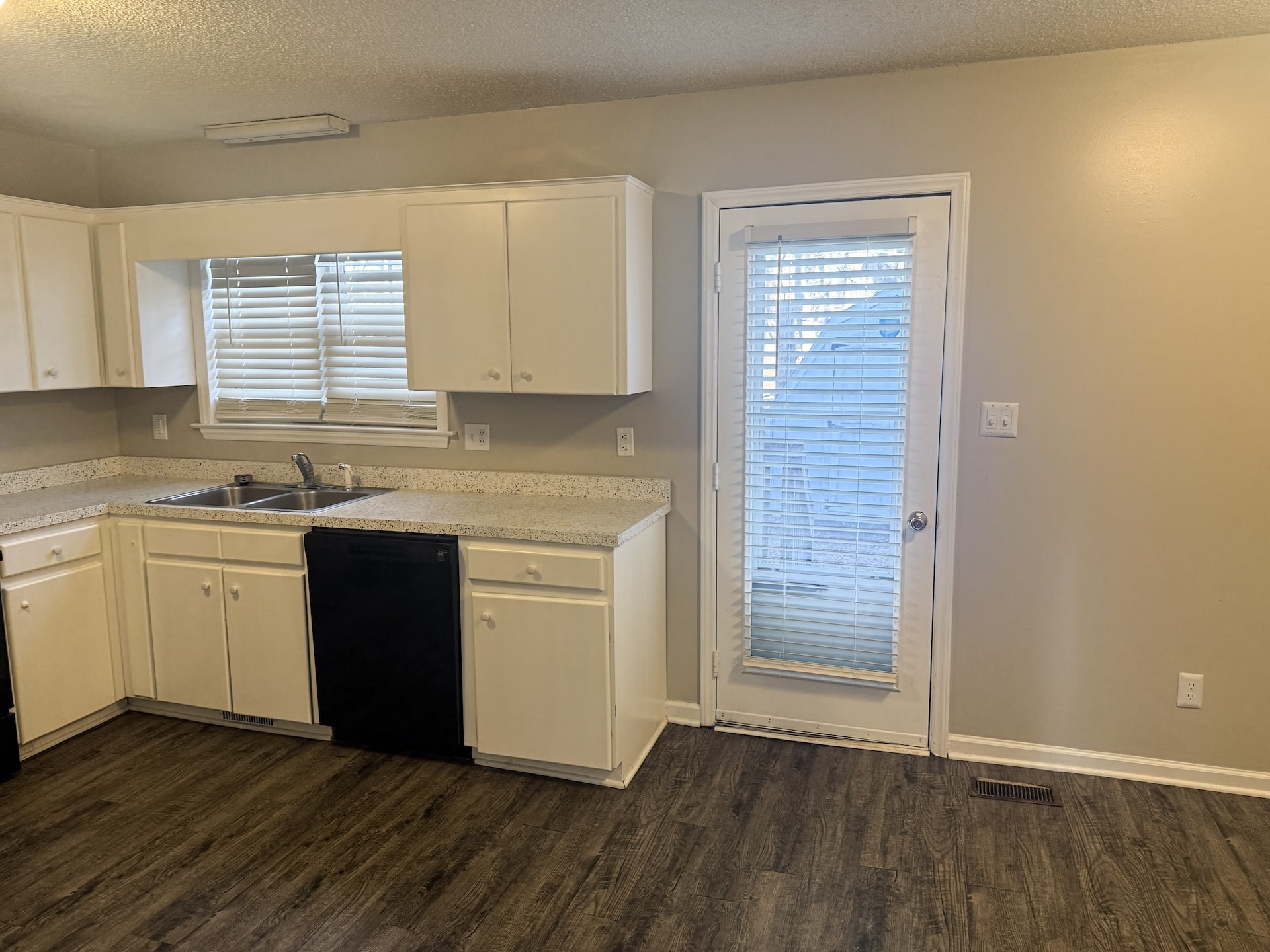 A kitchen with white cabinets, a double sink, and a black dishwasher. There's a window with blinds above the sink and a door with blinds leading outside, on a dark wood floor.