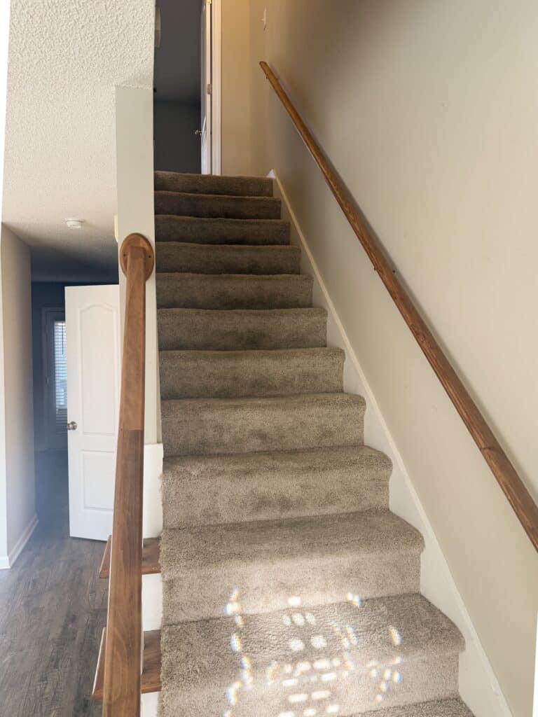 Carpeted stairs with a wooden handrail on the right, leading to a white door at the top. The wall is light-colored, and sunlight creates patterns on the steps.