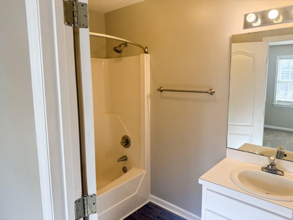 Bathroom with a white bathtub and shower curtain rod, a vanity with a sink and mirror, and an empty towel bar on a beige wall. Natural light enters through a window in the adjacent room.
